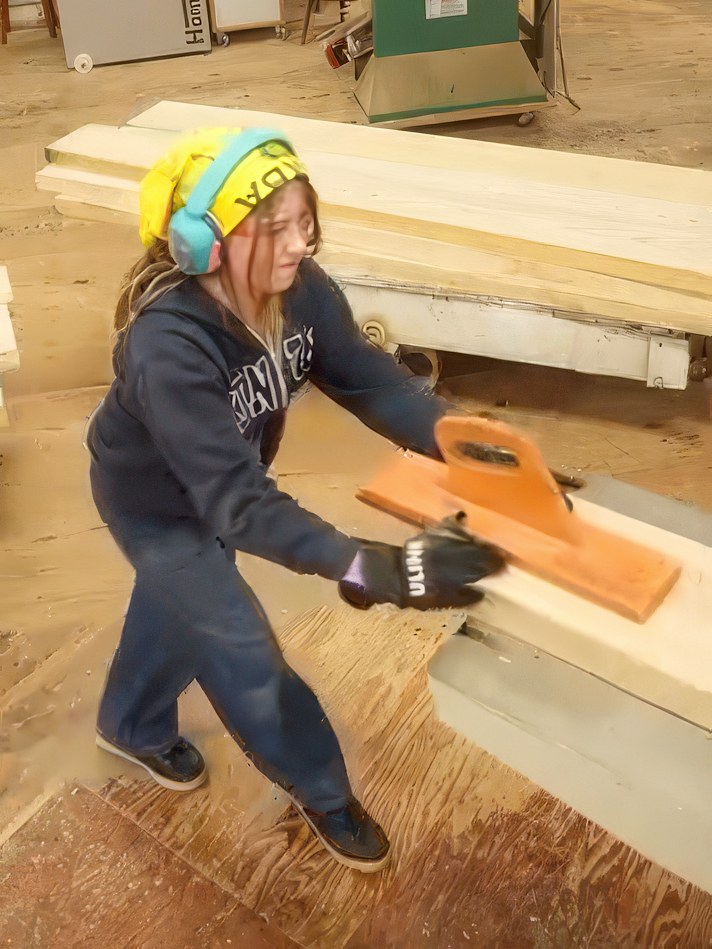 Willa feeding a board across the jointer with a push block, hearing protection on